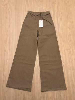 Bruna vida jeans från Avora - Säljer ett par bruna jeans från Avora, piazza trousers, med hög midja och vida ben. Jeansen har ett matchande bälte i midjan och klassiska fickor fram. De är aldrig använda och har prislappen kvar