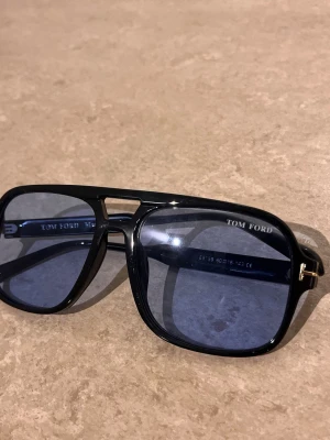 Svarta Tom Ford solglasögon - Snygga svarta solglasögon från Tom Ford med blåtonade glas och klassisk pilotform. Bågarna har gulddetaljer på skalmarna och en stilren design som ger en exklusiv känsla. Perfekta för dig som vill ha en trendig accessoar till sommaren.