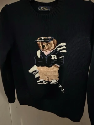 Svart stickad tröja Polo Ralph Lauren - Svart stickad tröja från Polo Ralph Lauren med ikonisk björn på bröstet. Tröjan har rund halsringning och långa ärmar. Björnen är broderad och bär collegekläder i blått, vitt och beige. Perfekt för dig som gillar klassisk preppy stil.