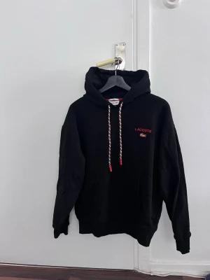 Svart hoodie från Lacoste - Svart hoodie från Lacoste med röd logga och krokodil på bröstet. Snygga snören med röd detalj och stor färgglad krokodil- och textdesign på ryggen. Perfekt för dig som gillar streetwear.