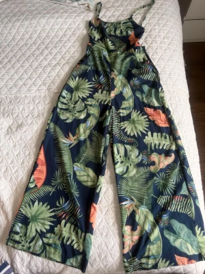 Blommig jumpsuit med vida ben - Säljer en snygg jumpsuit med tropiskt bladmönster i grönt, orange och blått. Modellen har smala axelband, markerad midja och vida ben för en avslappnad look. Perfekt för sommardagar och festivaler. Materialet känns lätt och luftigt.