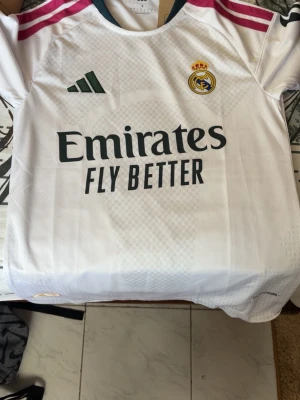 Real Madrid matchtröja Adidas vit - Snygg vit Real Madrid matchtröja från Adidas med klubbmärke på bröstet och sponsortryck 'Emirates Fly Better' framtill. Tröjan har rosa och mörkgröna detaljer på axlar och hals, samt korta ärmar. Tillverkad i lätt och ventilerande material, perfekt för fotboll.