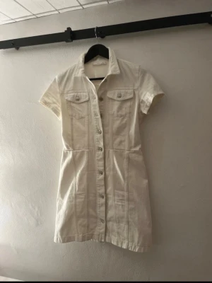 Vit kort jeansklänning med knappar - Snygg vit kort jeansklänning med knappar hela vägen framtill, krage och korta ärmar. Två bröstfickor med lock och markerad midja ger en cool och avslappnad look. Perfekt för dig som gillar stilrena och enkla plagg.
