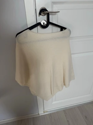 Beige poncho i mjukt tyg - En enkel och stilren beige poncho i tunt, mjukt material. Ponchon har en lös passform och faller snyggt över axlarna. Perfekt för att addera ett extra lager till din outfit och ge en avslappnad look.