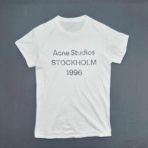 Vit Acne Studios t-shirt med tryck - Säljer en klassisk vit t-shirt från Acne Studios med svart texttryck på bröstet. T-shirten har rund hals och korta ärmar. Perfekt basplagg med stilren design och skön passform i mjuk bomull.