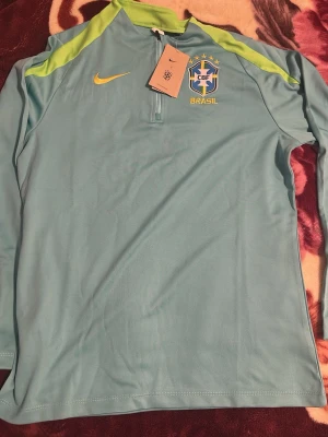 Nike Brasil långärmad träningströja - Ljusblå långärmad träningströja från Nike med Brasilien CBF-logga och gula detaljer. Tröjan har halv dragkedja framtill och limegröna axelpartier. Perfekt för fotbollsträning eller sportiga aktiviteter.