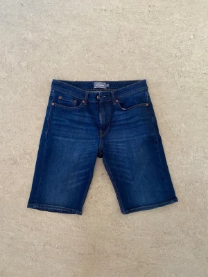 Mörkblå jeansshorts - Storlek S. Snygga mörkblå jeansshorts med klassisk femficksdesign och knappgylf. Shortsen har raka ben och bälteshällor i midjan. Perfekta för en avslappnad stil under varmare dagar.