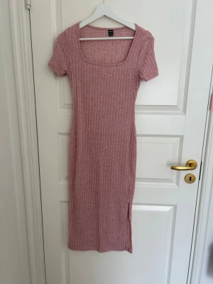 Rosa ribbad klänning med slits - Rosa ribbad klänning med korta ärmar och fyrkantig ringning. Klänningen är figurnära och har en slits på sidan för extra stilpoäng. Perfekt för dig som gillar en enkel men trendig look.