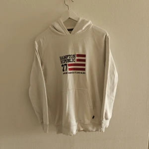 Hampton Republic hoodie - Vit hoodie från Hampton Republic med tryck och röda ränder framtill. Klassisk känguruficka, huva och ribbade muddar. Mjuk insida i bomullsblandning, perfekt för chill dagar. Snygg och enkel att matcha med jeans eller joggers.