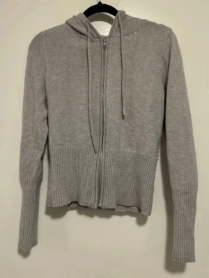 Ljusgrå stickad hoodie med dragkedja - Mysig ljusgrå hoodie i stickat material med dragkedja framtill och ribbade muddar vid ärmslut och nederkant. Utrustad med huva och dragsko för extra komfort. Perfekt för en avslappnad och stilren look.