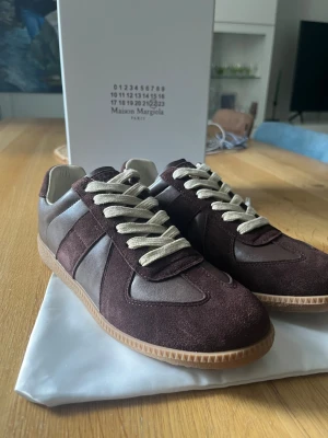 Maison Margiela bruna sneakers - Snygga sneakers från Maison Margiela i mörkbrunt mocka och läder med beigea skosnören och gummisula. Klassisk low-top siluett som ger en clean och stilren look. Perfekta för dig som gillar exklusiva och tidlösa sneakers.