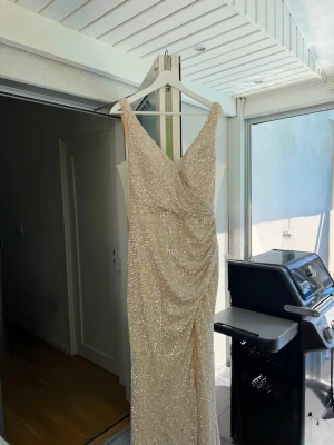 Champagnefärgad glittrig långklänning - Säljer en elegant långklänning i champagnefärg med glittriga paljetter över hela klänningen. Klänningen har v-ringning, åtsittande passform och  slits framtill. Nypris: 2445kr