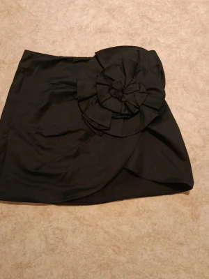 Svart kortkjol med stor rosett - Snygg svart kortkjol med en stor dekorativ rosett framtill. Kjolen har omlottkänsla och dragkedja bak. Perfekt för dig som vill sticka ut med en unik detalj. Passar till många olika stilar och tillfällen.