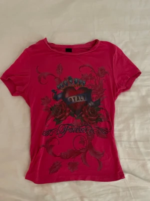 Rosa t-shirt med tryck och rosor - Y2k Cool rosa t-shirt med tryck av hjärta, krona och rosor samt texten 'Loyalty Forever' på framsidan. T-shirten har korta ärmar och en klassisk rund halsringning. Perfekt för dig som vill sticka ut med en unik och färgstark stil.