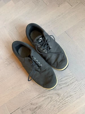 Nike Golfskor  - Göra golfs sneakers med låg profil och vita detaljer. Ovandelen är i textil och skon har svarta skosnören samt metallfärgade öljetter. Yttersulan är svart med gult rutmönster och vit Nike-logga. Perfekta för en sportig och avslappnad stil. Saknar innersula