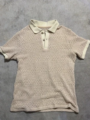 Beige stickad pikétröja - Snygg beige stickad pikétröja med kort ärm och klassisk krage. Tröjan har knappar framtill och är gjord i ett luftigt, virkat material som ger en cool retrovibe. Perfekt för dig som gillar en avslappnad men stilren look.