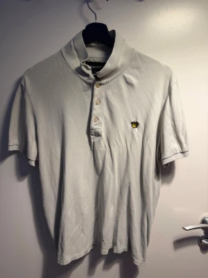 Vit pikétröja från Lyle & Scott - Hej säljer en Klassisk vit pikétröja från Lyle & Scott med korta ärmar och knappar framtill. Tröjan har det ikoniska gula logomärket broderat på bröstet och är tillverkad i mjuk bomull. Perfekt för en stilren och avslappnad look.