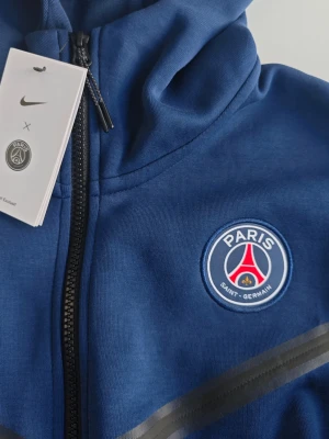 Blå PSG hoodie från Nike - Le Paris ! Ny snygg blå hoodie från Nike med Paris Saint-Germain-logga på bröstet och dragkedja framtill. Tröjan har huva, svarta detaljer och är tillverkad i ett mjukt funktionsmaterial. Perfekt för träning eller att bära till vardags om du gillar sportig stil.