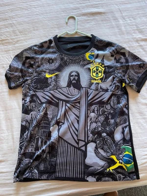  Brasilien jesus fotbollströja - Ruggigt snygg Brasilien tröja. Köpt på marknad i Spanien för 45 euro. Unik svart fotbollströja från Nike med Brasilien-tema. Tröjan har ett stort tryck av Kristusstatyn i Rio, detaljerade grafiska mönster och gula logotyper. På bröstet finns Brasiliens landslagsemblem och Nike-logga. Materialet är lätt och andas, perfekt för träning eller match.