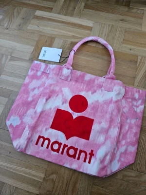 Rosa shoppingväska från Isabel Marant - Snygg tie dye shoppingväska i rosa och vitt batikmönster från Isabel Marant. Väskan har två rejäla handtag och ett stort rött logotryck på framsidan. Tillverkad i slitstarkt tyg, perfekt för att bära med sig allt man behöver. Supersnygg accessoar för dig som gillar färg!