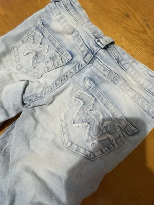 Ljusblå jeans med broderade bakfickor - Säljer ett par ljusblå jeans med snygga broderade detaljer på bakfickorna. Jeansen har klassisk femficksdesign och bälteshällor. Materialet är denim i bomull och de har en cool, tvättad look.