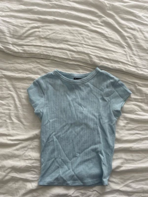 Ljusblå ribbad t-shirt - En ljusblå t-shirt med ribbad struktur och rund hals. T-shirten har korta ärmar och är gjord i ett mjukt material som passar perfekt till en avslappnad stil. Liten i storleken.