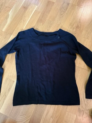 Svart långärmad tröja med knapp - En enkel svart långärmad tröja med rund hals och en dekorativ knapp vid axeln. Tröjan har en smal passform och är tillverkad i ett mjukt, tunt material som passar perfekt till olika outfits.