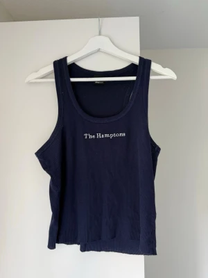 Marinblå ribbad linnetopp Gina Tricot - Snygg marinblå ribbad linnetopp från Gina Tricot med texten 'The Hamptons' broderad framtill. Toppen är ärmlös och har en avslappnad passform, perfekt till sommaren. Enkel att matcha med jeans eller shorts.