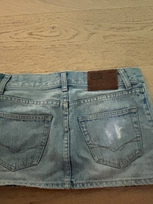 Kort jeanskjol från Levi's - Säljer en snygg kort jeanskjol från Levi's i ljusblå denim med slitna detaljer och klassiska bakfickor. Kjolen har bälteshällor och en brun logopatch bak. Perfekt för dig som gillar en avslappnad och trendig look.