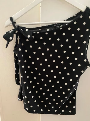 Svart prickig topp - Svart oneshoulder topp med vita prickar och en snygg knyt-detalj på axeln. Toppen är ärmlös och har en figurnära passform som ger en trendig look. Perfekt för dig som vill sticka ut lite extra med ett klassiskt mönster. Storleken är L men skulle säga att den är mer S