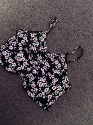 Blommig svart croppad linnetopp - Supersöt croppad linnetopp med smala axelband och svart botten täckt av blommigt mönster i vitt, rosa och grönt. Toppen har rynkad baksida för extra stretch och sitter tajt. Perfekt för sommarens alla outfits!