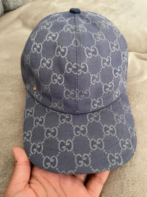 Blå GG-mönstrad keps från Gucci - Snygg blå keps från Gucci med det klassiska GG-mönstret över hela kepsen. Tillverkad i ett tåligt tygmaterial och har en böjd skärm. Perfekt accessoar för dig som vill ha en exklusiv och trendig look.