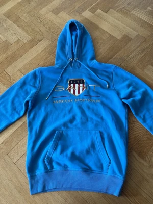 Blå hoodie från GANT med tryck - Snygg blå hoodie från GANT med broderad logga och texten 'American Sportswear' på bröstet. Klassisk känguruficka framtill och justerbar huva med dragsko. Perfekt för en avslappnad och sportig stil. Pris kan diskuteras!