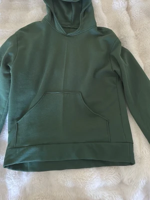 Grön hoodie  - En enkel och stilren grön hoodie med huva och stor magficka framtill. Hoodien har långa ärmar och är gjord i ett mjukt material som känns skönt mot huden. Perfekt för dig som gillar en clean look och vill ha något bekvämt att slänga på dig.