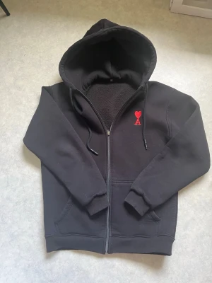 Svart hoodie med röd Ami-logo - Svart hoodie med dragkedja och huva från Ami. Den har ett rött hjärta med bokstaven A broderat på bröstet. Hoodien har ribbade muddar, fickor framtill och är gjord i mjukt, fleecefodrat material. Perfekt för dig som gillar stilrena detaljer och komfort.