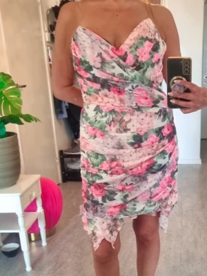 Blommig omlottklänning från ASOS - Supersnygg kort blommig klänning från ASOS med omlottdesign och volangdetaljer. Klänningen har en v-ringad urringning och är ärmlös. Färgerna går i vitt, rosa och grönt med ett romantiskt blommönster. Perfekt för sommarens alla tillfällen!