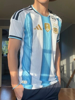 Argentina VM 2022 Champions Tröja – Adidas – Ny med tags - Säljer en mycket snygg Argentina-tröja med VM 2022 Champions-märke på bröstet. Tröjan är i nyskick och har fortfarande originaltags kvar. Perfekt för samlaren, Argentina-supportern eller dig som vill bära världsmästarnas ikoniska matchtröja.  • Argentina landslagströja • Adidas • FIFA World Champions 2022-märke • Tre stjärnor ovanför AFA-emblemet • Ny med tags kvar • Ventilerande ClimaCool-material • Mycket hög detaljnivå i tyg och tryck  Storlek: M (L men passar som M) Pris: 449 kr