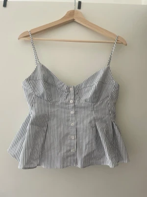 Randig linnetopp från Zara - Säljer en vit och blå smalrandig linnetopp från Zara med tunna axelband och knappar framtill. Toppen har en figurnära passform och lätt utsvängd nederdel, perfekt för varma dagar. Materialet känns svalt och mjukt mot huden.