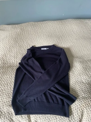 Mörkblå stickad tröja från Ryvls - Mörkblå stickad tröja från Ryvls med rund halsringning och långa ärmar. Tröjan har ribbade muddar vid ärmslut och nederkant, vilket ger en clean och klassisk look. Perfekt för lager på lager och enkel att matcha med olika outfits.