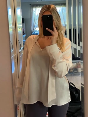 Ljusrosa satinblus från MQ - Snygg beige blus i satin från MQ med v-ringning och dekorativa band framtill. Blusen har en lös passform och långa ärmar 🩷