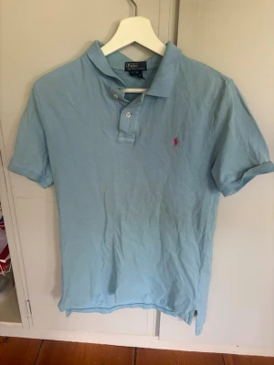 Ljusblå pikétröja från Polo Ralph Lauren - Klassisk ljusblå pikétröja från Polo Ralph Lauren med korta ärmar och krage. Tröjan har knappar vid halsen och den ikoniska lilla röda logotypen broderad på bröstet. Tillverkad i 100% bomull för en skön och luftig känsla. Säljes pga den är för liten för mig! Står att det är storlek L men passar med s-m