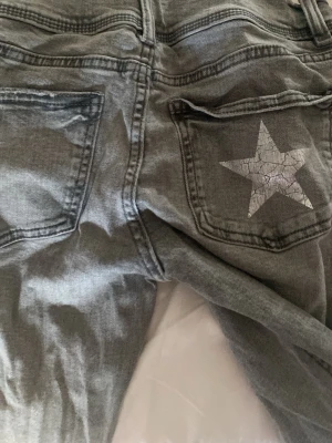 Grå jeans med stjärndetalj - Säljer ett par grå lågmidjaxd jeans med utsvängda ben och en cool  stjärna på ena bakfickan. Jeansen har klassisk femficksmodell och är tillverkade i denim. Perfekta för dig som vill sticka ut lite extra med en unik detalj på baksidan.