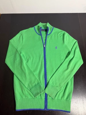 Grön ziptröja från Polo Ralph Laren  - Grön ziptröja från Polo Ralph Lauren med blå detaljer längs dragkedjan, kragen och nederkanten. Tröjan har en liten broderad logga på bröstet och är tillverkad i ett tunt, mjukt material. Perfekt för lager på lager och en färgklick i garderoben.