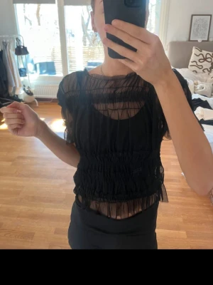 Svart transparent volangblus - Säljer en svart transparent blus med volangdetaljer och rynkade partier. Blusen har korta ärmar och är tillverkad i ett tunt, syntetiskt material som ger en luftig känsla. 