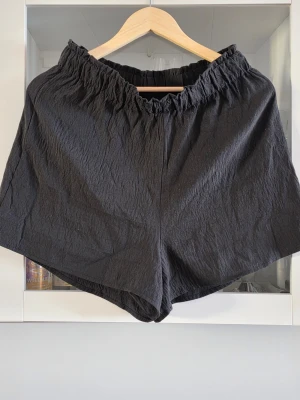 Svarta loose shorts med resårmidja - Säljer ett par svarta shorts med loose passform och bred resår i midjan. Materialet är lätt och har en skrynklig struktur som ger en avslappnad vibe. Perfekta för varma dagar och enkel att matcha med olika toppar.