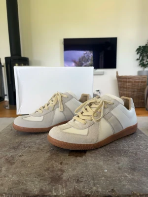 maison margiela gats vit - Stilrena sneakers i vit och ljusgrått med ovandel i läder och mocka. Skorna har klassisk design med låga skaft och bruna gummisulor som ger bra grepp. Perfekta för dig som gillar minimalistisk och tidlös stil. Boxen är lite trasig men påverkar ej skon