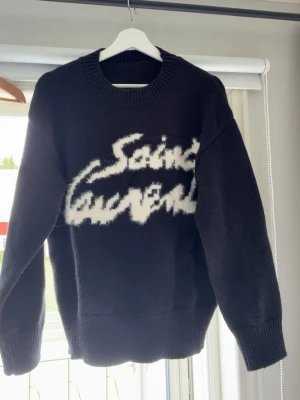 Svart Saint Laurent stickad tröja - Säljer en svart stickad tröja från Saint Laurent med vit, fluffig logotyp framtill. Tröjan har rund halsringning, långa ärmar och ribbade muddar vid ärmslut och nederkant. Perfekt för dig som gillar streetstyle och exklusiva märken.