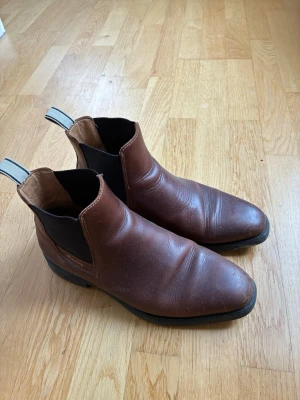Bruna Chelsea boots från Gant - Säljer ett par klassiska Chelsea boots från Gant i brunt läder med svarta resårpaneler på sidorna och robust svart sula. Bootsens dragflikar har vit och svart randig detalj. Perfekta för dig som gillar stilrena och tidlösa skor.