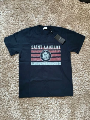 Svart Saint Laurent t-shirt med tryck - Snygg svart t-shirt från Saint Laurent med coolt tryck framtill i vitt och rött. T-shirten har rund halsringning och korta ärmar. Materialet är mjuk bomull och passformen är normal. Perfekt för dig som vill ha en trendig och stilren look.