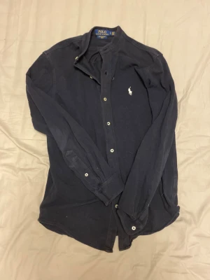 Mörkblå skjorta från Polo Ralph Lauren - Snygg mörkblå långärmad skjorta från Polo Ralph Lauren i featherweight mesh-material. Klassisk knappslå framtill och vit broderad logga på bröstet. Perfekt för dig som gillar stilrena och tidlösa plagg.